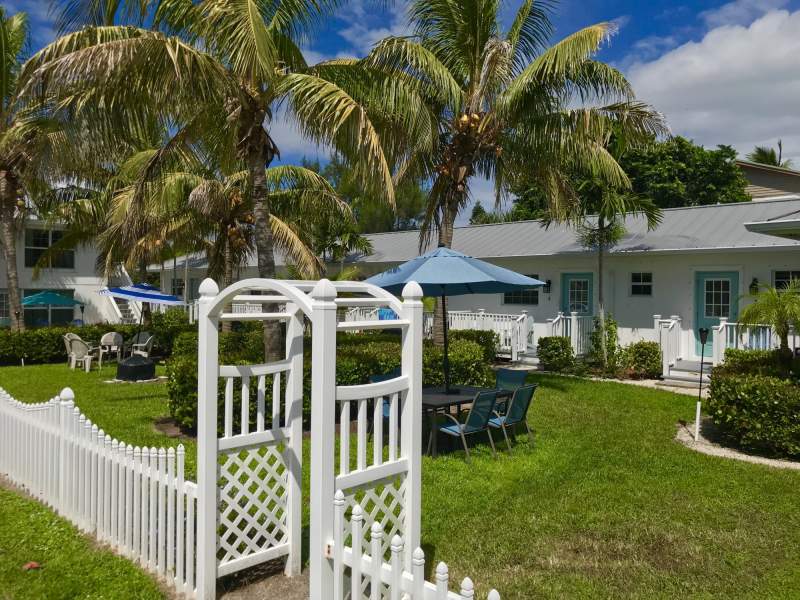 Exterior of bungalows, courtyard, tables, chairs, umbrellas, green grass, palm trees and white picket fence.