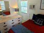 Double bed with maroon and blue bedding, white dresser with mirror, window with blinds, air conditioning unit