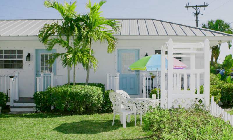 Exterior of bungalow, white building, metal roof, blue doors, palm trees, table, chairs and umbrella, trellis